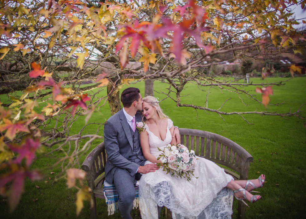 Groom kisses bride on forehead on a bench in the garden