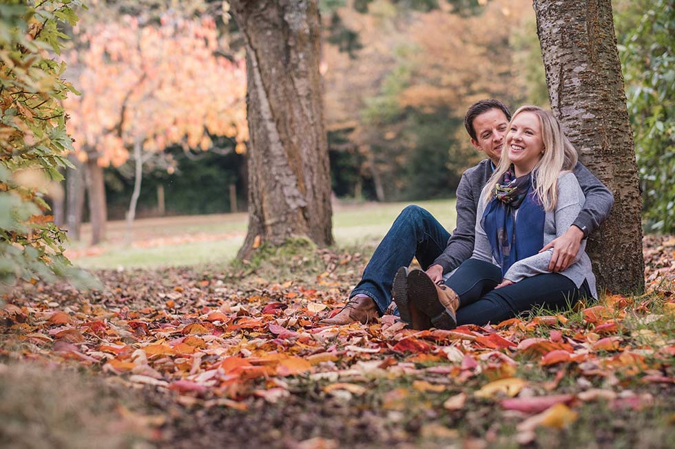 Bride and groom sitting at the tase of a tree amongst the autumn leaves at Wadhurst Castle
