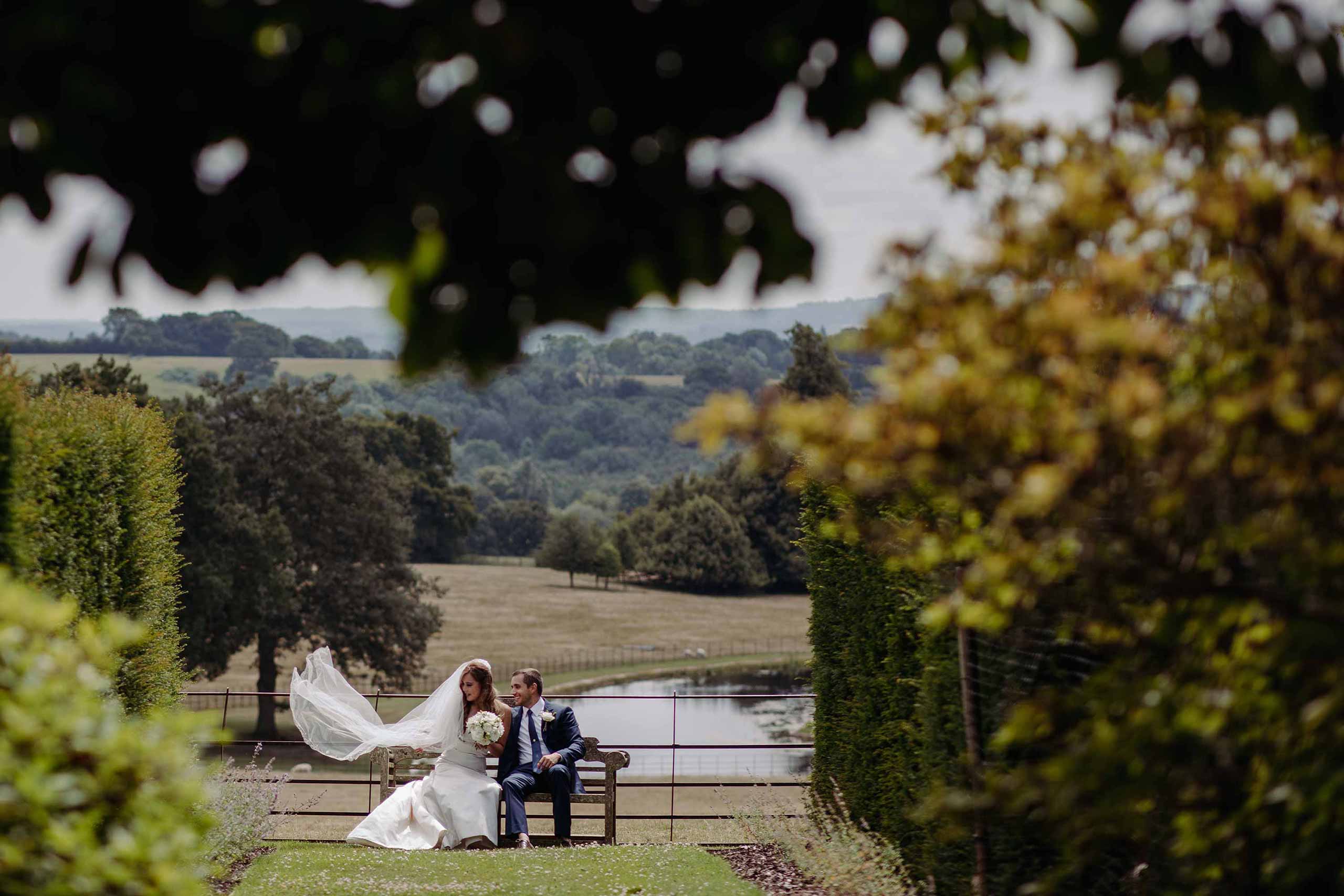 Bride and groom enjoying a quiet moment on a bench at Wadhurst Castle with the lake on view behind them