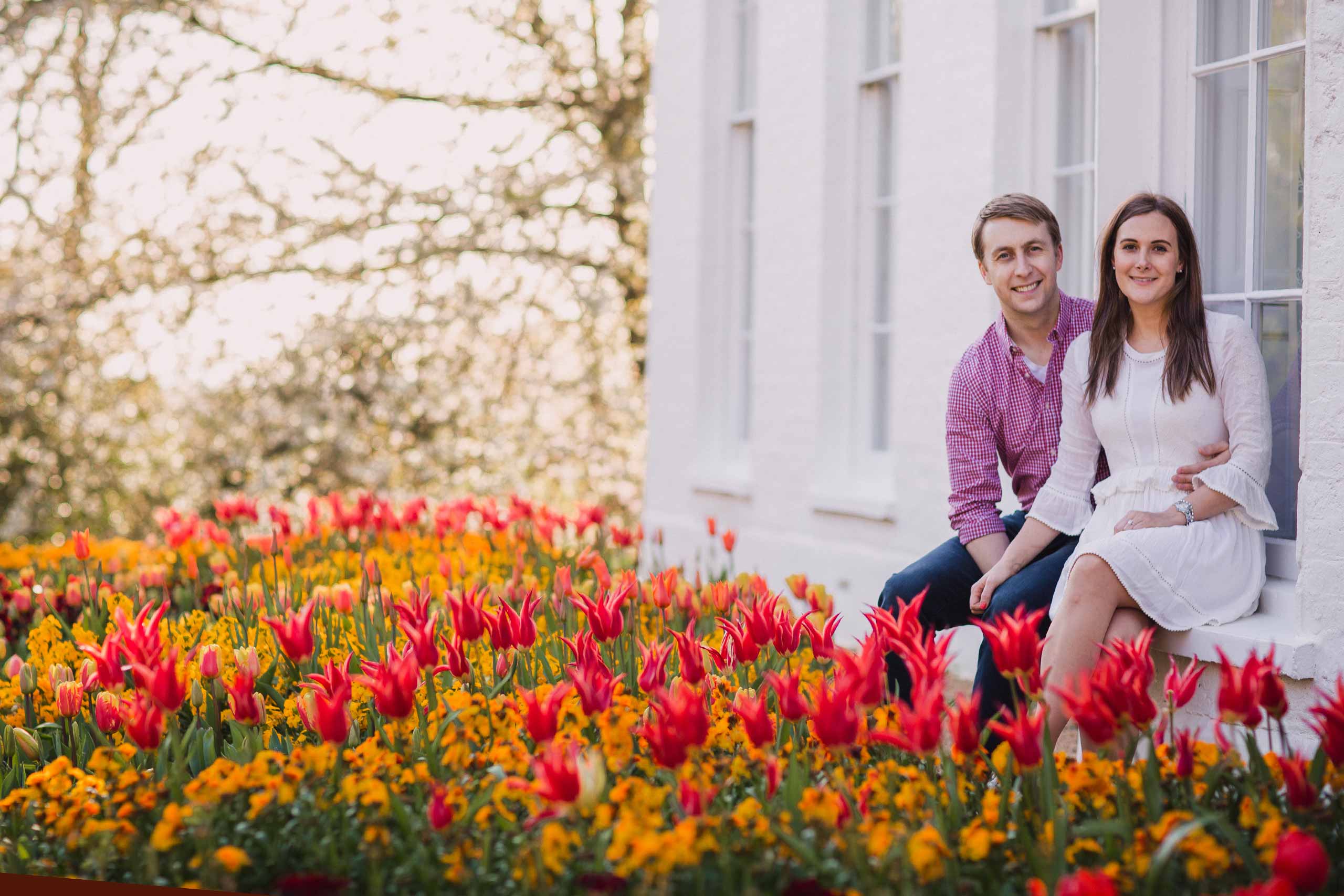 Bride and groom smiling at the camera whilst sitting on a window ledge above the tulips at Pembroke Lodge in Richmond Park