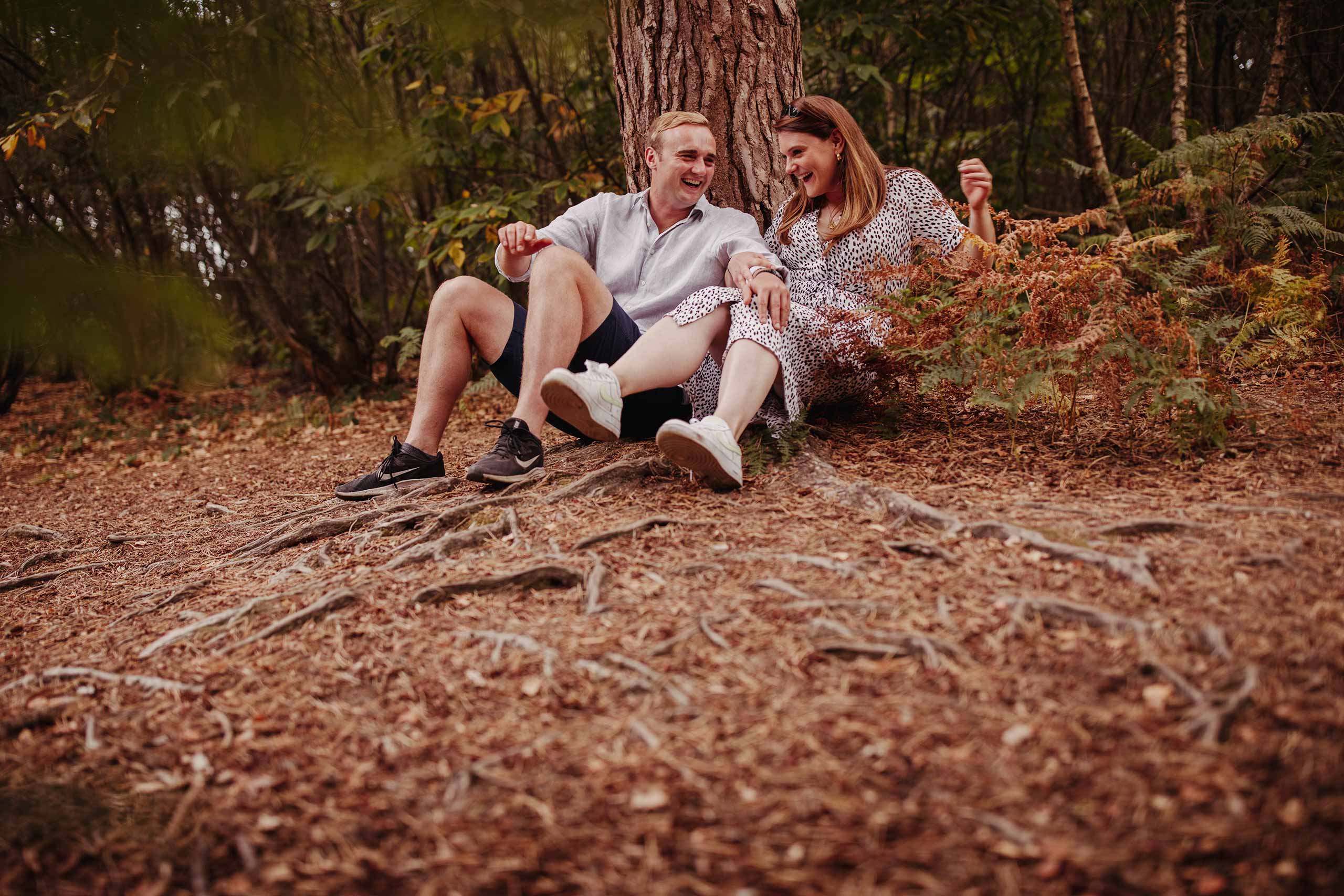Couple sitting against a tree laughing during their lockdown pre-wedding shoot in Sussex