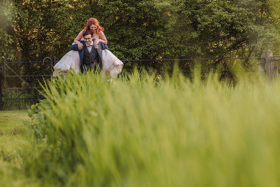 Groom gives bride a piggy back in the grass at Grittenham Barn