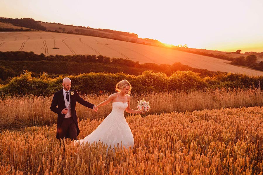 Bride and groom walk through a wheat field at sunset at Pangdean Barn