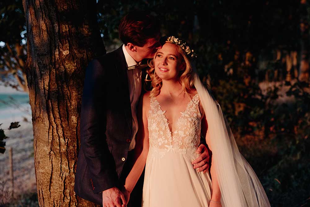 Groom kisses bride on the temple at golden hour in amongst the trees